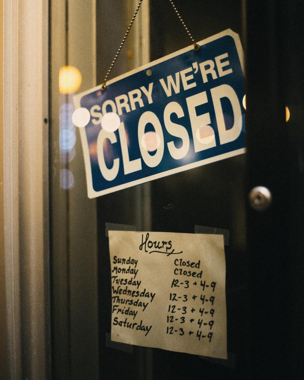 A sorry we're closed sign on a shop door, above a handwritten list of opening hours.