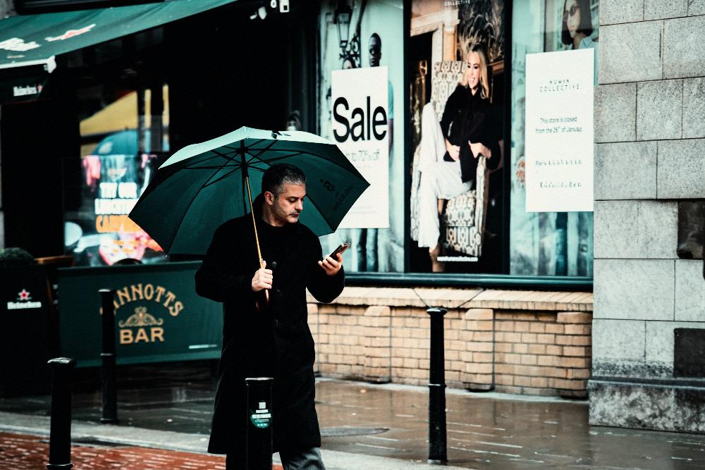 A man under an umbrella checks his phone on a wet pavement outside a shop.